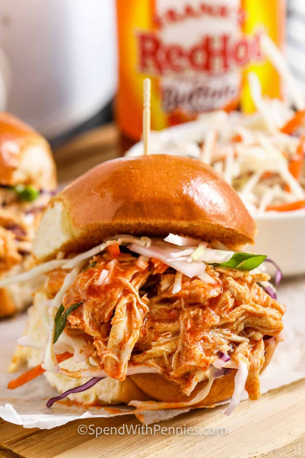 close up of Slow Cooker Buffalo Chicken sandwich with a bottle of buffalo hot sauce in the back