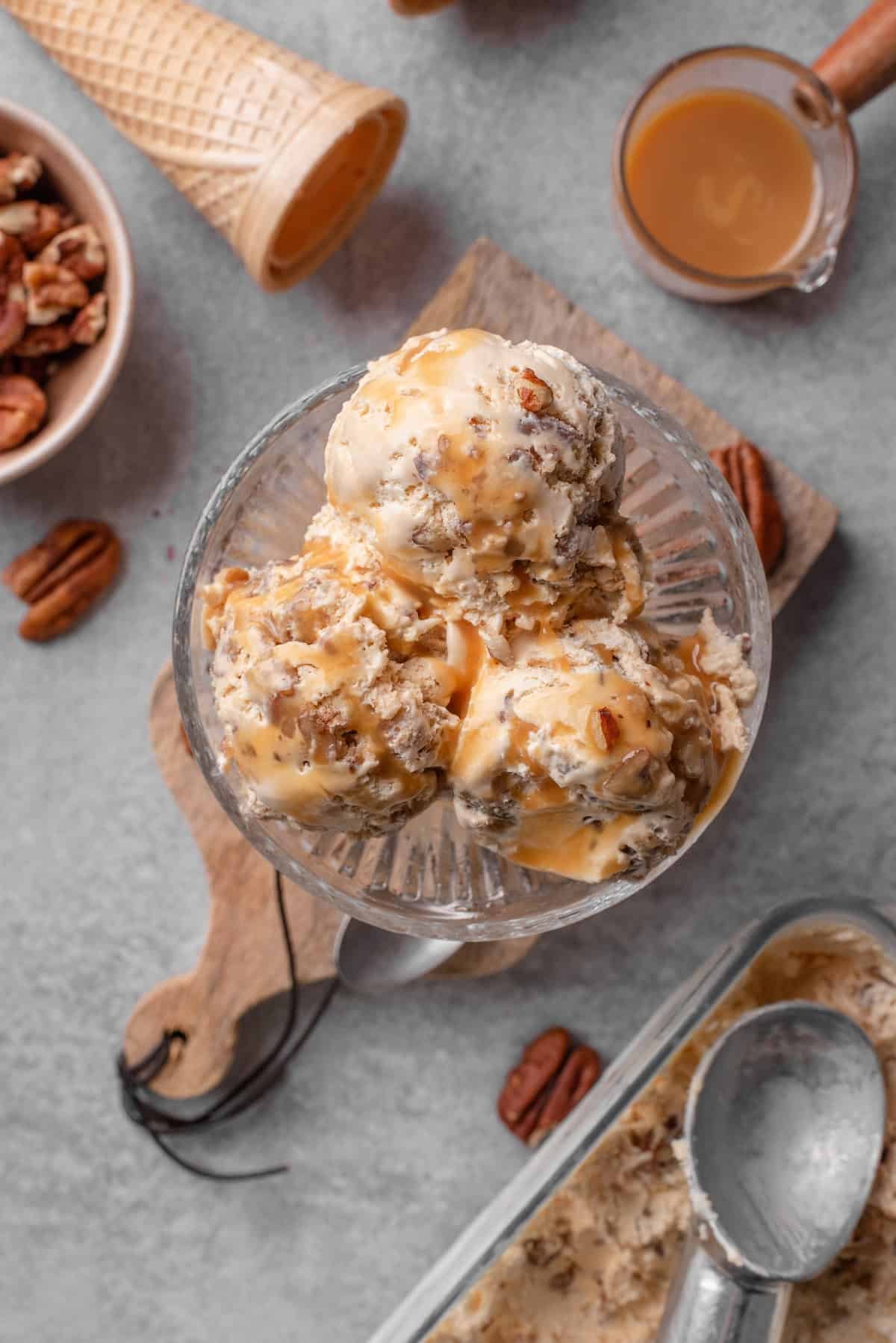 Overhead view of scoops of butter pecan ice cream in a glass dessert bowl, surrounded by ingredients.