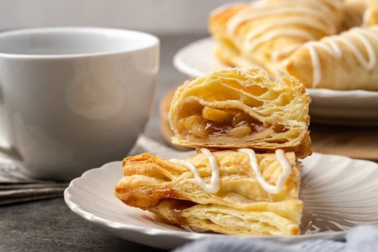Two halves of an apple turnover stacked on a plate with a coffee cup and more turnovers in the background.
