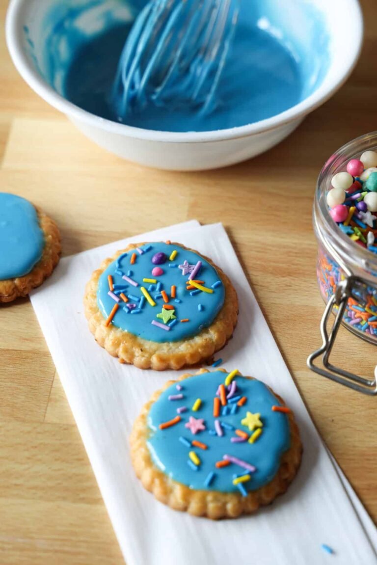 Two cookies decorated with blue sugar cookie icing and sprinkles on a countertop, with a bowl of blue icing in the background.
