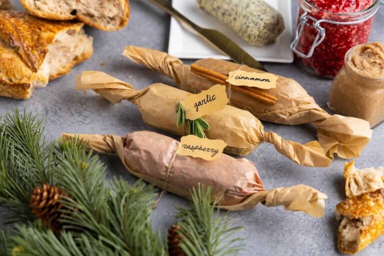 Close-up of wrapped compound butters labeled raspberry, garlic, and cinnamon on a gray background. Fresh raspberries and frosted holiday greenery are arranged around the butters, with jars of raspberry and cinnamon butter in the background.