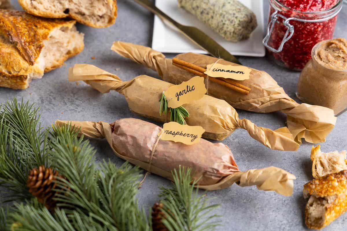 Close-up of wrapped compound butters labeled raspberry, garlic, and cinnamon on a gray background. Fresh raspberries and frosted holiday greenery are arranged around the butters, with jars of raspberry and cinnamon butter in the background.