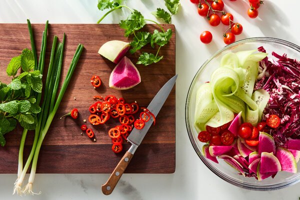 An overhead image of fruits and vegetables being sliced and added to a bowl.