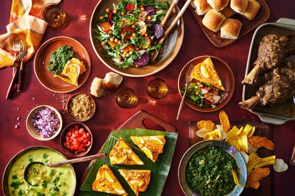 An overhead image of a festive feast featuring lamb shanks, moin moin, soup, salad and a bright green dip.