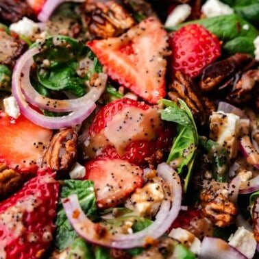 Closeup of strawberries, sliced red onions, pecans, and spinach, drizzled with a poppy seed dressing.