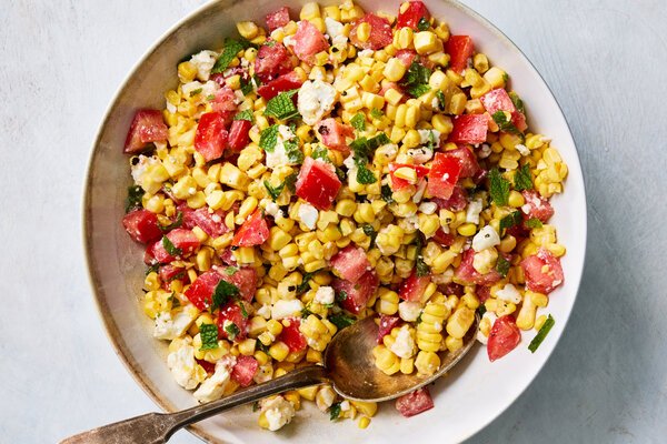 A vibrant salad in a bowl, with yellow corn, red tomatoes, white feta cheese and green mint. A spoon is partly visible.